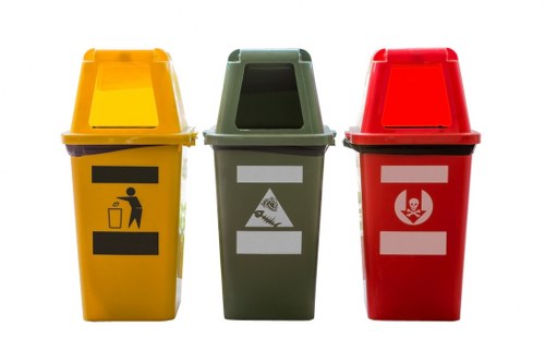 Three colour-coded waste disposal bins are positioned side by side outdoors on a paved surface, with a yellow bin on the left, a green bin in the middle, and a red bin on the right. The yellow bin features a black symbol of a person disposing of waste, indicating general rubbish; the green bin displays a white symbol of a pizza slice and a bottle, suggesting recyclable or organic waste; the red bin shows a white symbol of a biohazard or hazardous waste icon. The bins are made of plastic with smooth, slightly textured surfaces, and each has a matching lid that is slightly raised at the front, allowing for easy access. The scene appears evenly lit, with natural daylight suggesting an outdoor setting, possibly near a commercial or communal waste collection point. The environment emphasizes waste segregation and disposal practices, aligning with services provided by Business Waste Removal Mill Hill in the local area. The background is neutral, focusing entirely on the waste containers, with no additional objects or environmental features visible.