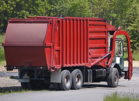 A large red waste collection truck parked on a paved surface in a rural or semi-rural area, with a lush green backdrop of trees and grass. The truck features a prominent, rectangular rear compaction container made of metal with visible vertical ridges and a smooth, curved top edge. The side of the container is partly open, revealing a clean, painted interior. To the right of the vehicle, a hydraulic lifting mechanism with a prominent arm and support strut is attached, designed for lifting and emptying large refuse bins. The truck's cab is white with a green stripe, and the vehicle is equipped with three axles and large black tires. This scene is representative of rubbish collection services, such as those provided by Business Waste Removal Mill Hill, operating in the local area around NW7 postal code, serving the Mill Hill community in London.