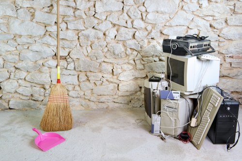 A collection of old electronic devices, including a CRT computer monitor, a tower CPU, a wired keyboard, and a black speaker, are stacked against a textured stone wall in what appears to be a garage or storage area. The devices are surrounded by tangled cables and are placed on a concrete floor. To the left, a broom with a wooden handle and a pink dustpan are positioned in front of the stone wall, which has a mix of irregularly shaped light-colored stones. The environment suggests an area used for disposing of or storing miscellaneous waste, with the electronic items indicating an electronics or household waste context. The scene is lit by natural or ambient light, highlighting the rough surface of the stone wall and the various textures of the electronics and cleaning tools. This setting aligns with the services offered by Business Waste Removal Mill Hill, focusing on rubbish removal, including obsolete electrical appliances.