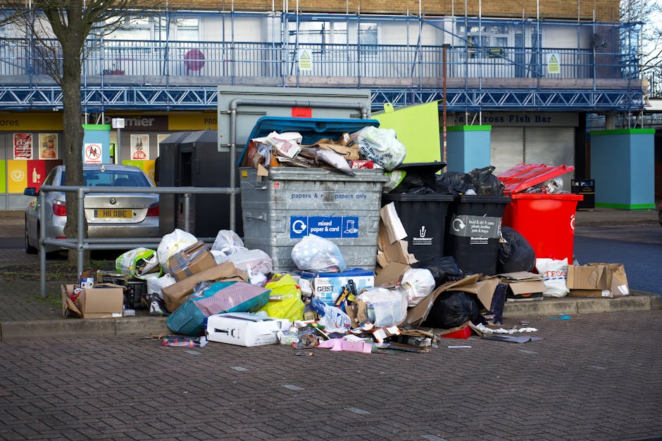 An overflowing collection of various types of waste and rubbish bags is piled in front of multiple bins on an urban street, with some cardboard boxes and flattened packaging scattered around the base.