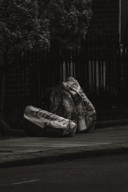 An overflowing collection of various types of waste and rubbish bags is piled in front of multiple bins on an urban street, with some cardboard boxes and flattened packaging scattered around the base.
